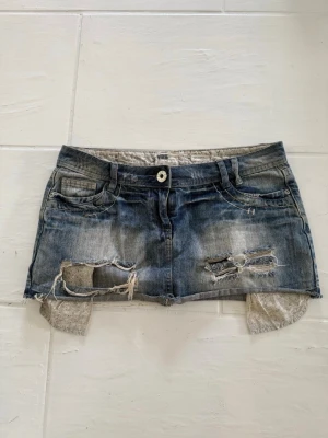 Kort blå jeanskjol med spets - Kort jeanskjol i blå tvättad denim med slitna detaljer och råa kanter. Kjolen har fickor fram och bak samt fina fickor med blommönster som sticker ut under nederkanten för en unik och trendig look. Perfekt för sommaren!!🩷.        Kolla gärna in mina andra annonser, bundle går att ordna för bättre pris!!