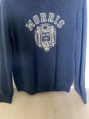 Blå stickad tröja från Morris - Mörkblå stickad tröja från Morris med vitt universitetsinspirerat tryck och emblem på bröstet. Tröjan har rund halsringning, långa ärmar och ribbade muddar vid ärmslut och nederkant. Perfekt för en avslappnad men snygg stil.