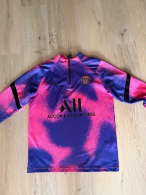 PSG träningströja lila/rosa - Säljer en långärmad träningströja från PSG i lila och rosa med svart logga och dragkedja vid halsen. Tröjan har ett coolt mönster och är tillverkad i ett lätt och snabbtorkande material, perfekt för fotbollsträning eller gym.