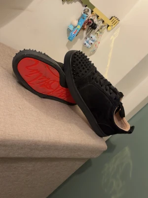 Svarta Louboutin sneakers med nitar - Säljer ett par svarta sneakers från Christian Louboutin med mattsvarta nitar på tån och klassisk röd sula med logga. Skorna har snörning och är tillverkade i mocka och läder för en lyxig vibe. Perfekta för dig som vill sticka ut med edgy detaljer.