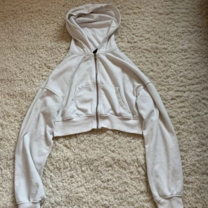 Vit croppad hoodie med dragkedja - Säljer en vit croppad hoodie med dragkedja framtill och två fickor. Hoodien har långa ärmar, ribbade muddar och en klassisk huva. Perfekt för en avslappnad och trendig stil.