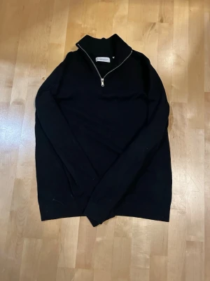 Svart halvzip stickad tröja TeeShoppen - Snygg svart stickad tröja från TeeShoppen med halvzip och hög krage. Tröjan har långa ärmar och ribbade detaljer vid kragen. Perfekt för lager på lager och enkel att matcha med olika outfits.