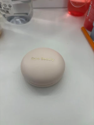 Rare Beauty rosa puderrouge - Säljer ett puderrouge från Rare Beauty i en mjuk rosa nyans. Kommer i en rund, ljusbeige dosa med spegel i locket och elegant guldtext. Perfekt för att ge kinderna en naturlig glow och fräsch färg.