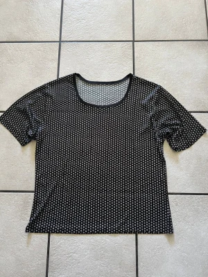 Svart topp med vita prickar storlek M/L - Trendig svart  topp med vita prickar och rund halsringning. Toppen är kortärmad och har en lätt transparent känsla som ger en cool vibe. Perfekt att styla lager på lager eller bära som den är för en edgy look.