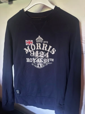 Mörkblå sweatshirt från Morris - Snygg mörkblå sweatshirt från Morris med tryck framtill i vitt och en liten brittisk flagga. Tröjan har rund halsringning och långa ärmar. Tillverkad i mjukt bomullsmaterial och har en klassisk, avslappnad passform. Perfekt för en casual och stilren look.