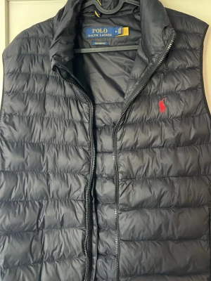 Svart dunväst från Polo Ralph Lauren - Snygg svart dunväst från Polo Ralph Lauren i storlek M med dragkedja framtill. Perfekt att slänga över en hoodie eller tröja när det är kyligt ute. Västen är i nästan nytt skick! Skriv vid funderingar👌🏼