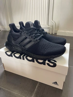 Svarta Adidas Ultraboost sneakers - Snygga helsvarta Adidas Ultraboost sneakers med stickad ovandel i mesh och klassiska tre ränder på sidorna. Skorna har en svart Boost-sula för extra komfort och sportig look. Perfekta för dig som gillar stilrena och bekväma sneakers.