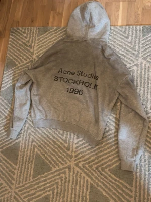 Grå hoodie från Acne Studios - Snygg grå hoodie från Acne Studios med tryck på ryggen där det står 'Acne Studios STOCKHOLM 1996'. Klassisk passform med huva och ribbade muddar vid ärmar och nederkant. Tillverkad i mjukt bomullsmaterial för en bekväm känsla. Endast provad aldrig brukat. Står S men passar som M eller baggy S.