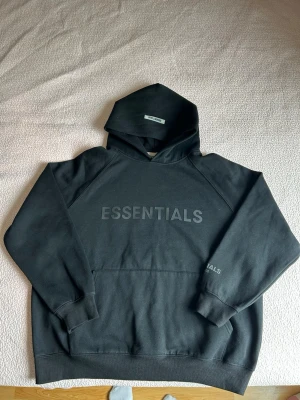 Svart Essentials hoodie med huva - Snygg svart hoodie från Essentials med stor logga framtill. Klassisk passform med huva och känguruficka. Tillverkad i mjukt material som känns skönt mot huden. Perfekt för en avslappnad streetwear-look.
