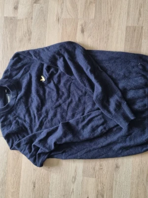 Mörkblå stickad tröja från Lyle & Scott - Mörkblå stickad tröja från Lyle & Scott med rund halsringning och den klassiska gula logon broderad på bröstet. Tröjan har långa ärmar och ribbade muddar vid ärmslut och nederkant. Perfekt för en stilren och avslappnad look.
