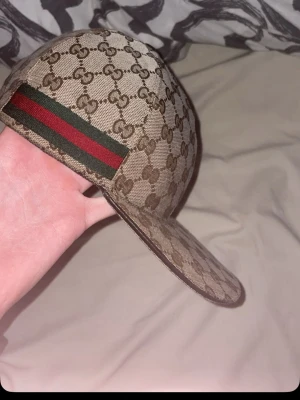 Gucci keps beige (kvitto, box finns ) - Säljer en beige keps från Gucci med klassiskt GG-monogram över hela kepsen och den ikoniska gröna och röda randen på sidan. Kepsen har böjd skärm och är tillverkad i canvasmaterial. Perfekt för dig som vill ha en lyxig och trendig accessoar.