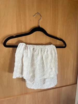 Vit kort kjol med broderade detaljer - Supersöt vit kort kjol med broderade blommor och volang. Kjolen har en bekväm resår i midjan och knytband framtill för extra passform. 