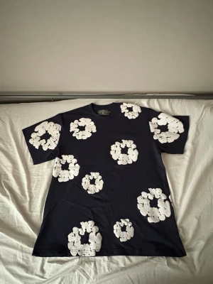  t-shirt från Denim Tears - Säljer en mörkblå t-shirt från Denim Tears med stora vita blommor tryckta över hela tröjan. T shirten är gjord i mjuk bomull. Perfekt för dig som vill sticka ut med ett unikt mönster.