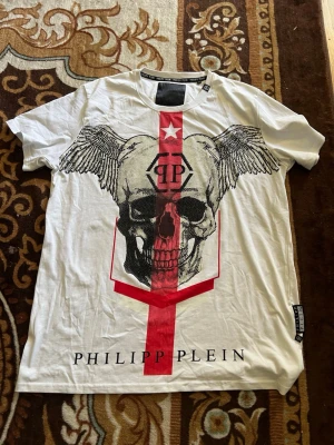 Vit Philipp Plein t-shirt med dödskalle - Snygg vit t-shirt från Philipp Plein med stort tryck av en dödskalle med vingar och röd vertikal linje framtill. T-shirten har korta ärmar, rund halsringning och exklusiv PP-logga på ryggen. Perfekt för dig som gillar statement-plagg.