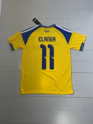 Sverige matchtröja Elanga #11 Adidas - Gul och blå Sverige matchtröja från Adidas med Elanga och nummer 11 på ryggen. Tröjan har korta ärmar, rund hals och klassiska Adidas-ränder på axlarna. Materialet är lätt och andas, perfekt för fotboll eller träning. Sveriges landslagsemblem på bröstet.