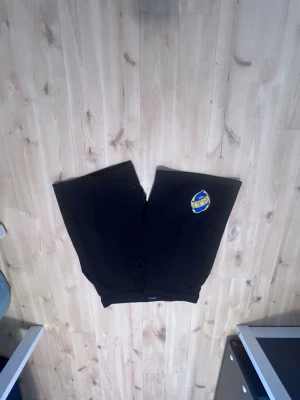 Svarta shorts med AIK-märke - Svarta shorts med AIK:s klubbmärke på vänster ben. Klassisk rak passform med resår i midjan och en bakficka. Perfekta för dig som vill representera AIK på ett avslappnat sätt. Tillverkade i mjuk bomull för skön känsla.
