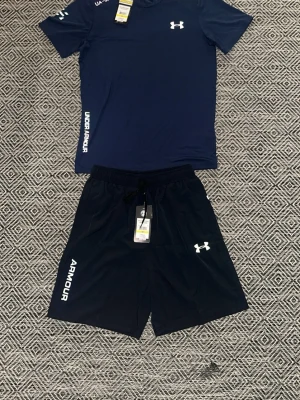 Blått träningsset från Under Armour - Snyggt träningsset från Under Armour med en mörkblå t-shirt och matchande shorts. Båda plaggen har logga och text i vitt. Tillverkade i ett lätt och snabbtorkande material som passar perfekt till gymmet eller löprundan. Storlek M men passar även S