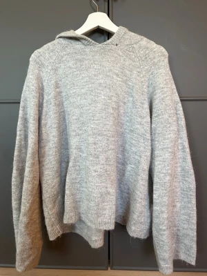 Grå stickad hoodie med luva - Mysig grå stickad hoodie med luva och långa ärmar. Tröjan har en relaxed passform och är tillverkad i ett mjukt stickat material som passar perfekt för kyliga dagar. Enkel och stilren design som är lätt att matcha med olika outfits.