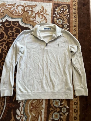 Ljusgrå quarter zip från Polo Ralph Lauren - Snygg ljusgrå långärmad tröja från Polo Ralph Lauren med klassisk quarter zip och broderad logga på bröstet. Tillverkad i mjukt bomullsmaterial och har ribbade muddar vid ärmslut och nederkant. Perfekt för en clean och avslappnad stil.