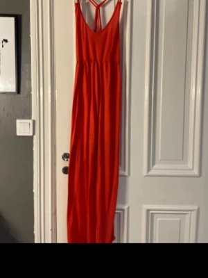 Röd långklänning med smala axelband - Säljer en röd långklänning med smala axelband och snygg ryggdetalj. Klänningen har en enkel, rak passform och är tillverkad i ett lätt material som passar perfekt till varma dagar. Stilren och trendig design.
