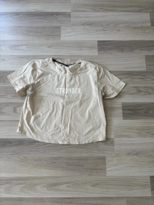 Beige t-shirt från Stronger - Snygg beige t-shirt från Stronger med vit text på bröstet. Tillverkad i mjuk och skön polyesterblandning med elastan för extra stretch. Klassisk rund halsringning och korta ärmar, lite croppad. Perfekt för en avslappnad och sportig look.