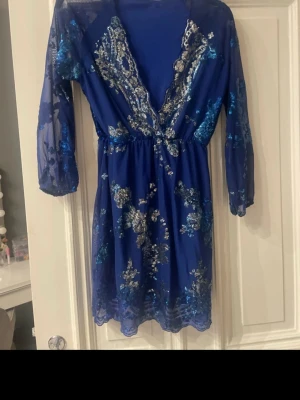 Blå glittrig kortklänning med spets - En blå kortklänning med transparenta ärmar och v-ringad spetsdetalj framtill. Klänningen har glittriga blommönster och är tillverkad i syntetmaterial. Perfekt för dig som vill sticka ut med en elegant look.