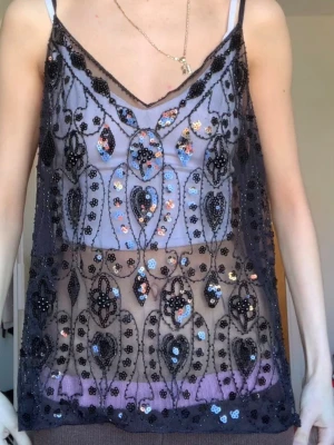 Svart mesh linnetopp med paljetter - Säljer en transparent svart mesh linnetopp med v-ringning och tunna axelband. Toppen är dekorerad med broderade detaljer och glittriga paljetter i svart, blått och rosa. Perfekt för dig som vill sticka ut med en unik och trendig look.
