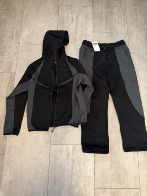 Nike svart och grå loungewear set - Ny pris ca 2500kr Nike loungewear set med zip-hoodie och byxor i svart och mörkgrått. Hoodien har dragkedja, huva och fickor, medan byxorna har dragsko i midjan och sidofickor. Båda plaggen har diskreta Nike-loggor och är tillverkade i mjukt, bekvämt material. Koftan är i storlek M men passar som S och byxan är storlek L men passar som M. Dem är en storlek mindre.