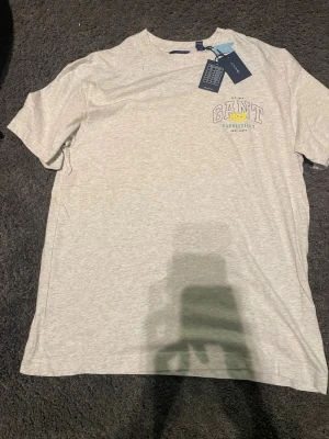 Ljusgrå GANT t-shirt med tryck  - Aldrig använd org pris 400 kr Snygg ljusgrå t-shirt från GANT med tryck på bröstet i vitt, gult och blått. Klassisk rund hals och korta ärmar. Tillverkad i mjuk bomull för skön känsla. Perfekt till jeans eller shorts för en clean och enkel stil.