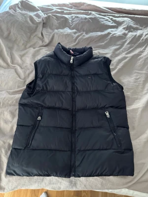 Svart dunväst från Tommy Hilfiger - Snygg svart dunväst från Tommy Hilfiger. 1 av innerfickorna jag ett hål i sig inuti. 