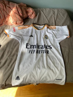 Real Madrid matchtröja Vini Jr. 7 - Säljer en vit Real Madrid matchtröja från Adidas med Vini Jr. och nummer 7 på ryggen. Tröjan har gula och svarta detaljer, La Liga-märke på ärmen och 'Emirates Fly Better' tryckt på bröstet. Tillverkad i lätt och ventilerande material, perfekt för fotboll.