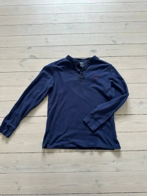 Mörkblå långärmad tröja Polo Ralph Lauren - Mörkblå långärmad tröja från Polo Ralph Lauren med liten röd logga broderad på bröstet. Tröjan har knappar vid halsen och är gjord i mjuk bomull. Enkel och stilren design som passar till det mesta. 10-12 år
