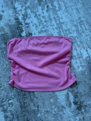 Rosa ribbad bandeau tubtopp - Säljer en rosa bandeau tubtopp med ribbad struktur. Toppen är axelbandslös och har en enkel, stilren design som passar perfekt till sommaren. Superenkel att matcha med olika outfits!