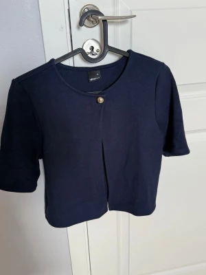 Mörkblå kortärmad kofta Gina Tricot - Stilren mörkblå kofta från Gina Tricot med korta ärmar och rund halsringning. Koftan har en enkel knäppning med en dekorativ knapp framtill och är gjord i ett mjukt, stretchigt material. Perfekt att slänga över en topp för en clean look.