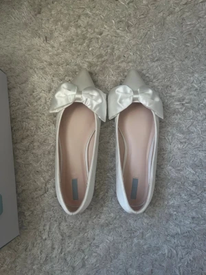 Vita ballerinaskor med rosett - Säljer ett par vita ballerinaskor med spetsig tå och stora, glansiga rosetter framtill. Skorna har en elegant och stilren design med slät yta och ljusrosa innersula. Perfekta för dig som gillar en söt och chic look. ALDRIG ANVÄNDA