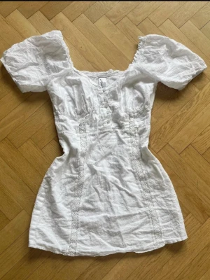 Vit kort klänning med puffärm - Söt vit kortklänning med puffärmar och spetsdetaljer. Klänningen har en v-ringad urringning och markerad midja som ger en feminin siluett. Tillverkad i ett lätt bomullstyg, perfekt för varma dagar. Passar dig som gillar romantisk och trendig stil. Storlek XS/S, fint skick!