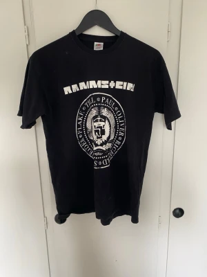 Svart Rammstein t-shirt med tryck - Cool svart t-shirt från Fruit of the Loom med stort vitt Rammstein-tryck på bröstet och bandmedlemmarnas namn runt en logga. Klassisk rund hals och korta ärmar. Köpt på en Rammstein konsert! Pruta om så vill😄