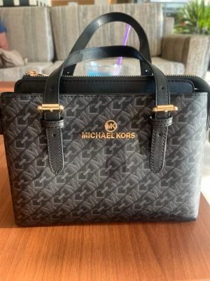 Svart handväska från Michael Kors - Snygg svart handväska från Michael Kors med guldiga detaljer och MK-logga framtill. Väskan har två handtag, dragkedja upptill och är täckt av ett diskret MK-mönster. Tillverkad i tåligt konstläder med stilren, boxig form. AXEL RÄM FÖLJES MED!