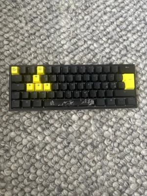 Mekaniskt tangentbord svart/gult - Snyggt kompakt mekaniskt tangentbord med svart bas och flera gula tangenter. Tangentbordet har unika tryck på vissa tangenter och konst på mellanslagstangenten. Undersidan är vit med justerbara fötter. Perfekt för gaming eller skrivande. Svarta swithes