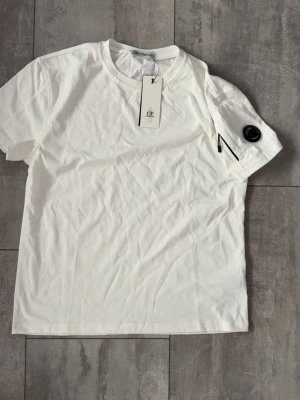 Vit t-shirt från C.P. Company - Snygg vit t-shirt från C.P. Company med klassisk rund hals och korta ärmar. På vänster ärm finns en ikonisk svart linsdetalj och en liten ficka med dragkedja. Tillverkad i mjuk bomull för skön känsla och stilren look.