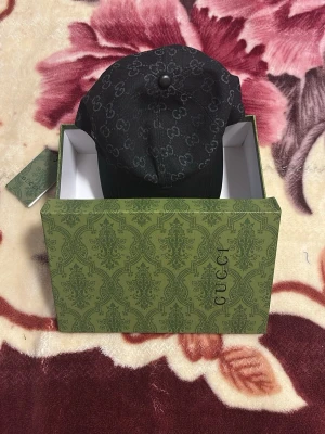 Svart keps från Gucci med monogram - Snygg svart keps från Gucci med klassiskt GG-monogram över hela tyget och böjd skärm i läderlook. Kepsen har en stilren design och levereras i original grön Gucci-box med mönster. Perfekt för dig som vill ha en exklusiv accessoar.