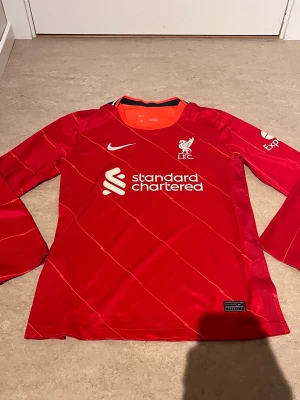 Liverpool långärmad matchtröja Nike - Röd långärmad Liverpool FC matchtröja från Nike med vita detaljer och klubbmärke på bröstet. Tröjan har tryckt sponsorlogga 'Standard Chartered' och tunna diagonala linjer över hela plagget. Tillverkad i funktionsmaterial som passar perfekt till träning eller match.