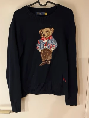 Svart stickad tröja Polo Bear - Stickad svart tröja från Polo Ralph Lauren med ikoniska Polo Bear-motivet framtill. Tröjan har rund hals, långa ärmar och ribbade muddar. RL-broderi i rött vid nederkanten. Perfekt för dig som gillar klassisk men lekfull stil. Har ett hål på baksidan i axeln som syns på bild.