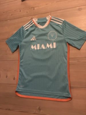 Inter Miami Messi matchtröja - Säljer en ljusblå Inter Miami matchtröja från Adidas med vita och orange detaljer. Tröjan har korta ärmar, rund hals och tryck med Messi och nummer 10 på ryggen. Materialet är lätt och andas, perfekt för fotboll eller träning.