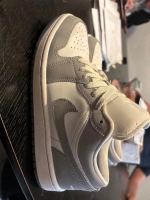 Nike Air Jordan 1 Low grå/vit - Snygga Nike Air Jordan 1 Low sneakers i grått och vitt läder. Klassisk siluett med perforerad tåbox, vita skosnören och ikoniska swoosh-loggan på sidan. Perfekta för dig som gillar stilrena och tidlösa sneakers.storlek 38,5
