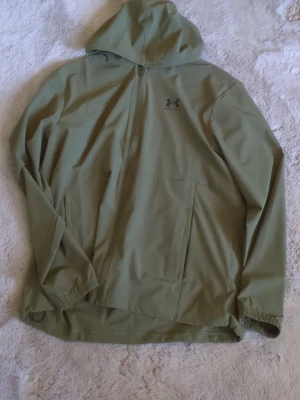Olivgrön vindjacka från Under Armour - Snygg olivgrön vindjacka från Under Armour med huva och dragkedja framtill. Jackan har två sidofickor och är tillverkad i ett lätt, vattenavvisande material. Perfekt för blåsiga dagar och sportiga outfits.
