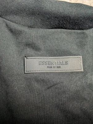 Svart Essentials Fear of God t-shirt - Snygg svart t-shirt från Fear of God Essentials med stor logga på ryggen och diskret patch vid nacken. Klassisk passform och tillverkad i mjuk bomull som känns skön mot huden. Perfekt för dig som gillar streetwear och stilrena plagg.
