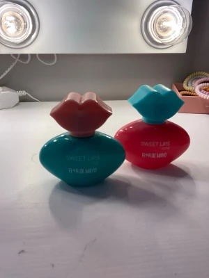 Sweet Lips parfym från Flor de Mayo - Två flaskor parfym från Flor de Mayo, Sweet Lips-serien. Den ena flaskan är turkos med ett rosa lock format som läppar, den andra är korallröd med ett turkost läppformad lock. Flaskorna har en lekfull och unik design i plast. 10 kr styck!💗 