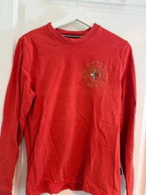 Röd långärmad topp från Tommy Hilfiger - Röd långärmad topp från Tommy Hilfiger med rund halsringning och broderad logga på bröstet. Tillverkad i mjuk bomull som känns skön mot huden. Perfekt för dig som gillar klassisk stil med en twist.