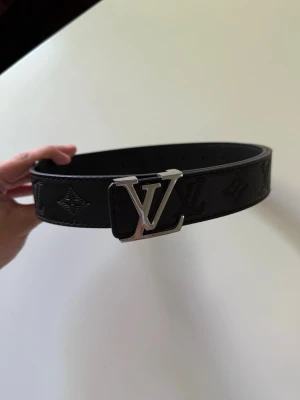 Svart Louis Vuitton skärp i skinn - Snyggt svart skärp från Louis Vuitton i skinn med klassiskt LV-spänne i silver och präglade monogramdetaljer längs hela bältet. Perfekt accessoar för att lyfta vilken outfit som helst.